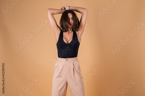 Fashionable portrait of a young woman on a beige background in a black bodysuit and trousers with gold cuts, a chain, a ring, posing