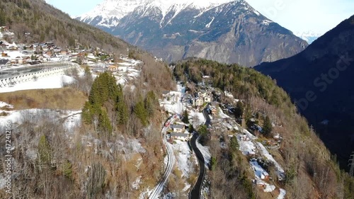 Aerial: red and white train in the Alps, going along a mountainside partially covered by snow and entering an alpine village photo