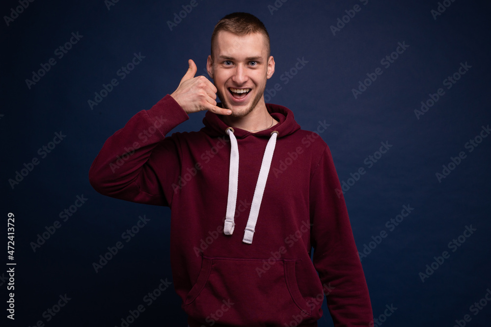 Young man in red hoodie and jeans posing on blue background and showing call gesture