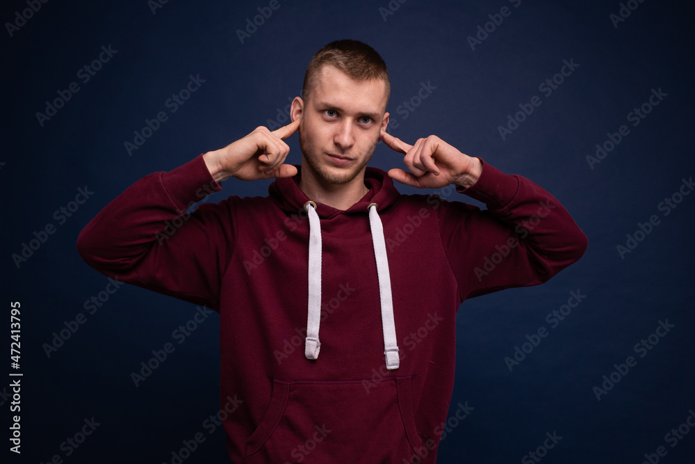 Young man in red hoodie and jeans posing on a blue background, covering his ears with his fingers