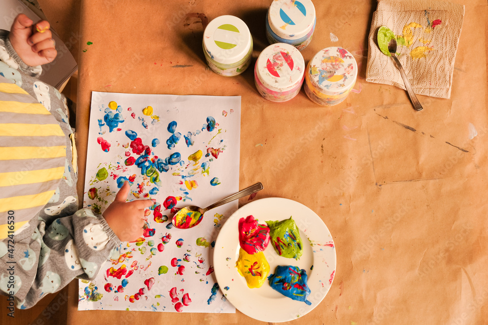 A child’s hand painting with bright finger paints at table covered with brown paper. Shot directly above A child’s hand painting with bright finger paints at table covered with brown paper. Shot directly above