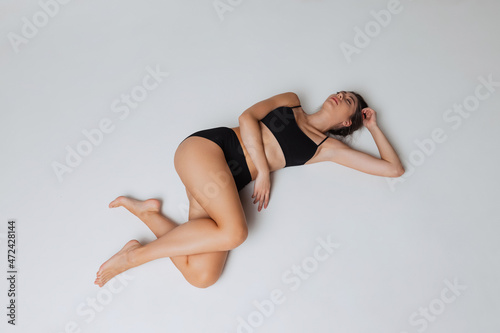 High angle view of young attractive slim girl in black underwear lying on floor isolated over gray studio background. Natural beauty concept.