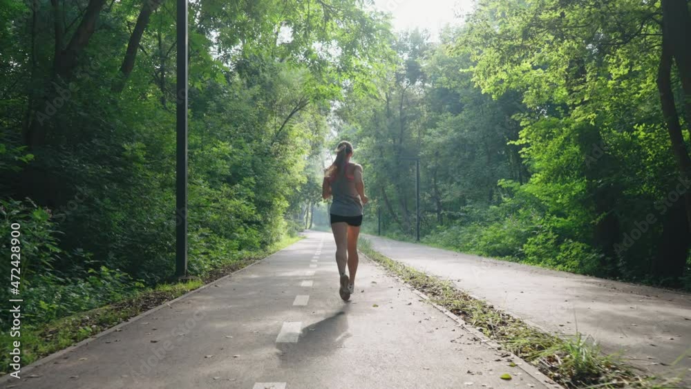 Female jogger wearing top and shorts running on alley in park in slow motion, sun shining through green foliage. Following shot fit woman exercising outside. Concept of healthy lifestyle
