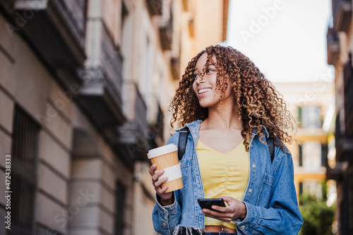 Beautiful woman looking away while holding reusable cup and smart phone in city photo