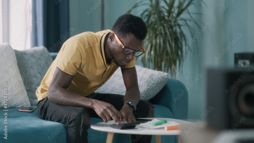 African American guy in glasses calculating utility bills and taxes and getting upset while sitting on sofa at home African American guy in glasses calculating utility bills and taxes and getting upset while sitting on sofa at home