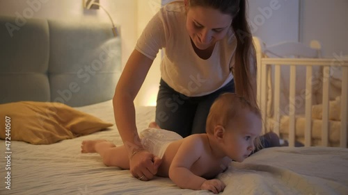 Caring mother putting on diapers on her little baby son crawling on bed at night. Concept of children hygiene, healthcare and family care at home