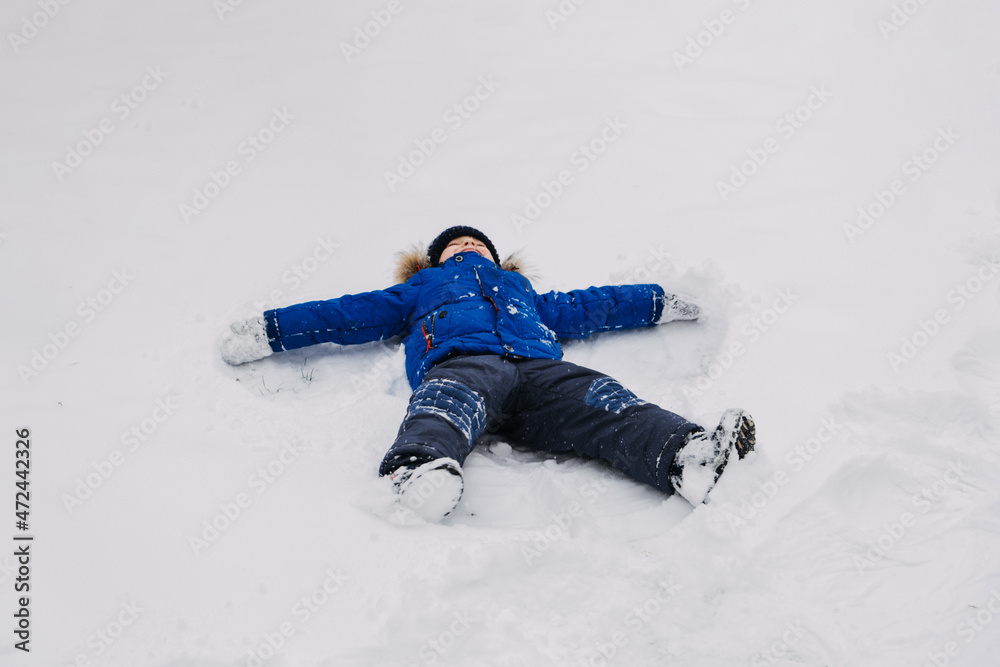 Best Outdoor Winter Activities for kids. Little kid boy in blue winter jacket making snow angel, laying down on snow in winter backyard. Active outdoors leisure with children in winter. Best Outdoor Winter Activities for kids. Little kid boy in blue winter jacket making snow angel, laying down on snow in winter backyard. Active outdoors leisure with children in winter.
