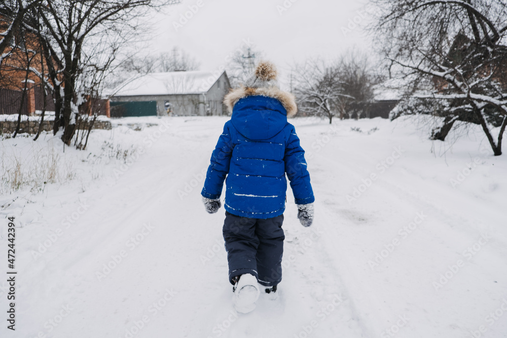 Best Outdoor Winter Activities for kids. Back view kid boy in blue winter jacket walking down a snowy street. Active outdoors leisure with children in winter.