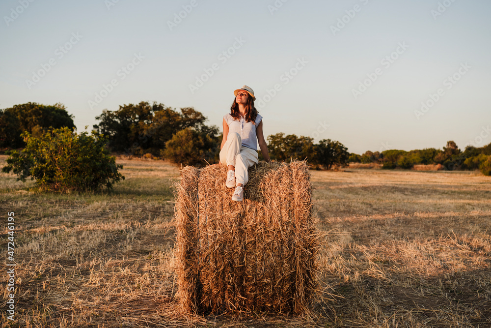 Woman wearing hat sitting on straw bale during sunset Woman wearing hat sitting on straw bale during sunset