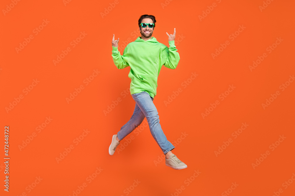 Full size photo of brunet young guy run show rock sign wear hoodie jeans sneakers eyewear isolated on orange background