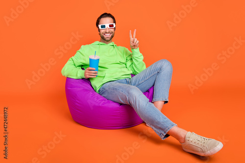 Full size photo of brunet millennial guy watch film drink show v-sign wear spectacles sweatshirt jeans shoes isolated on orange background