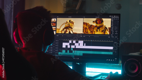 Teenager using computer to edit footage while sitting at desk in obscure room at night at home