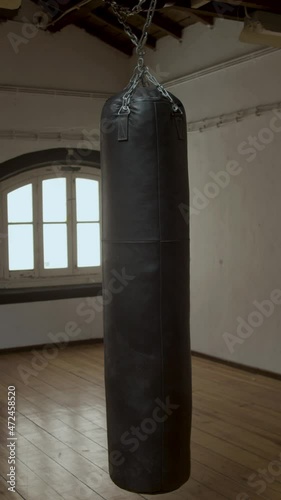 Vertical shot of swinging brown punching bag in empty gym. Slow motion of boxing room with windows in daytime. Sport, fitness, workout concept photo