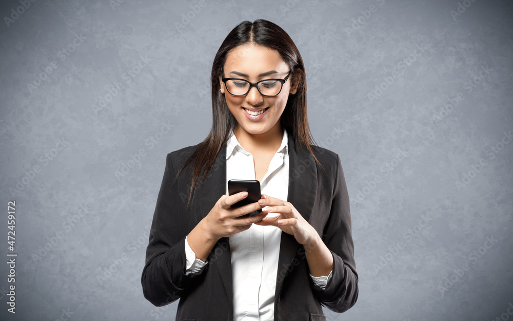 Studio shot of smiling young businesswoman in office wear with eyeglasses, holding smart phone standing on grey background Studio shot of smiling young businesswoman in office wear with eyeglasses, holding smart phone standing on grey background