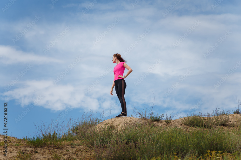 Young woman in sportswear standing on hill top on blue sky background. Fitness, healthy way of life, wellbeing, freedom, mental health, triumph, winning concept. Young woman in sportswear standing on hill top on blue sky background. Fitness, healthy way of life, wellbeing, freedom, mental health, triumph, winning concept.