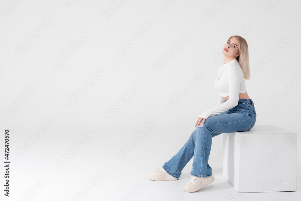 A young blonde in a white sweater and jeans sits and poses on an isolated white background in the studio. People lifestyle concept. Copy space for copy A young blonde in a white sweater and jeans sits and poses on an isolated white background in the studio. People lifestyle concept. Copy space for copy