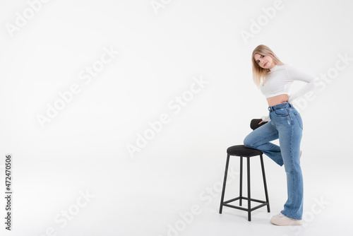 Happy and beautiful young woman in a white sweater and jeans with a black bar stool on a white background. People lifestyle concept.