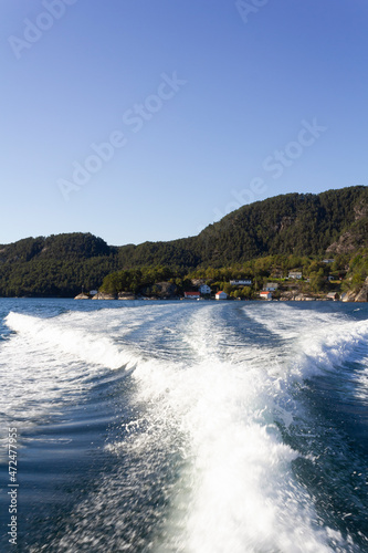 Trail on water surface of fast moving boat with norwegian green forest mountains on the background