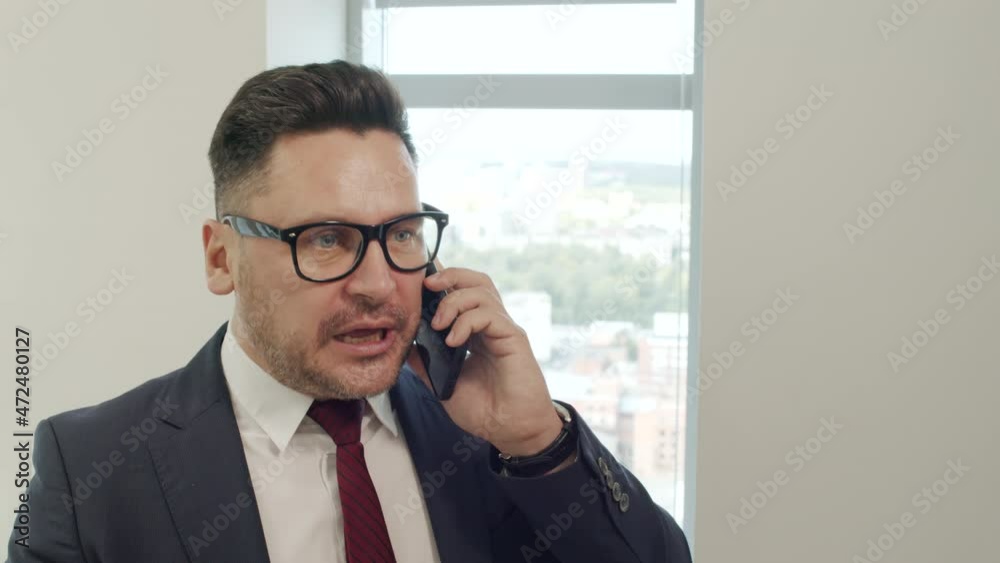Tracking close up shot of angry businessman in suit and glasses standing by window in office and having conversation with someone on mobile phone. He is talking and gesticulating Tracking close up shot of angry businessman in suit and glasses standing by window in office and having conversation with someone on mobile phone. He is talking and gesticulating