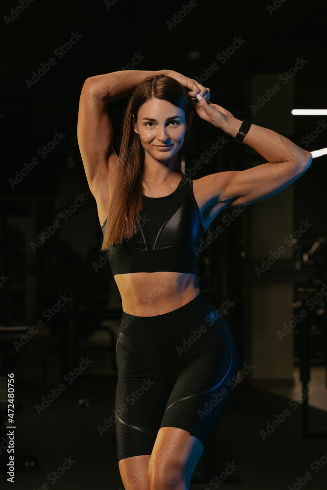 A fit girl in a black suit is posing in a gym. A sporty girl is stretching after a workout.