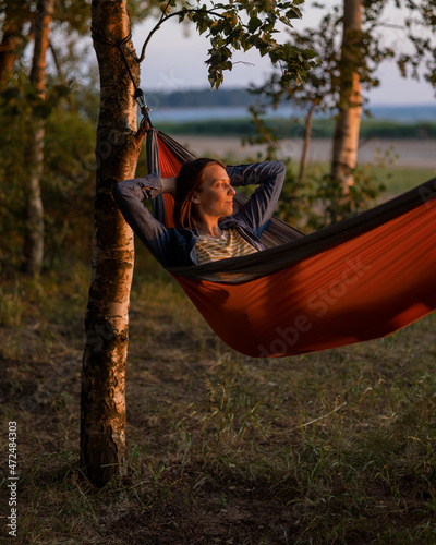 a European woman in a hammock in nature is resting and watching the sunset. a female travel blogger creates video content. hiking and outdoor recreation by the sea or in the woods. a cozy hammock