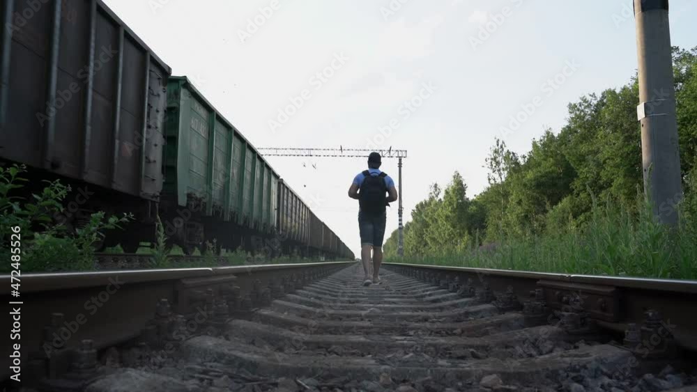 authentic male traveler with backpack walks distance along rails of railway near passing train on summer day authentic male traveler with backpack walks distance along rails of railway near passing train on summer day