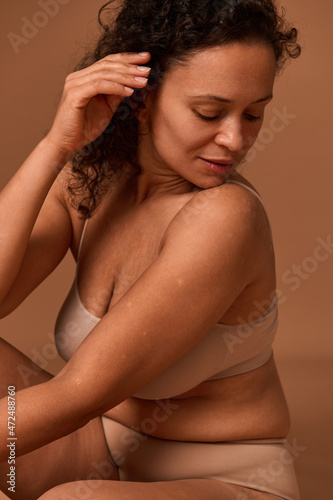 Portrait of a dark-haired curly woman with skin flaws, cellulite and stretch marks posing in beige underwear, looking down against colored background with copy space. Body positivity concept