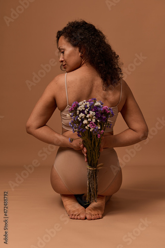 Rear view of beautiful African woman in lingerie, looking to the side, holding a bouquet of wildflowers behind her back, sitting on beige background. Body positive, self-acceptance, femininity concept