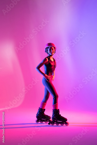 child athlete skater posing with arms on waist training with safety equipment for competition, looking at photo, on pink background in a studio