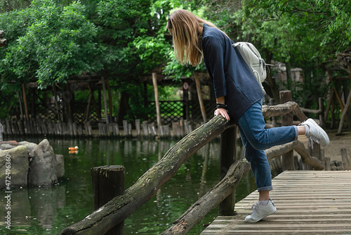 A girl bent over to an artificial pond in a public park and looks into the water from a wooden bridge. she has a white backpack on her shoulders. Travel and excursions alone, enjoying nature. © Ольга Рязанцева