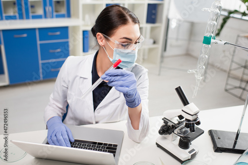 Scientist in latex gloves holding pcr test and using laptop near microscope in lab.