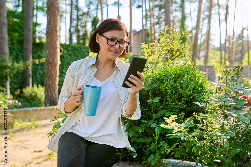 Fototapeta premium Middle-aged woman having rest in spring garden with cup of tea and smartphone in hands