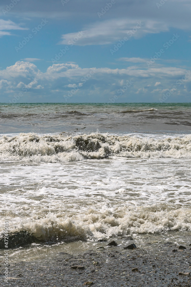 stormy waves of the Mediterranean sea run over the rocky summer beach against the backdrop of a blue cloudy sky