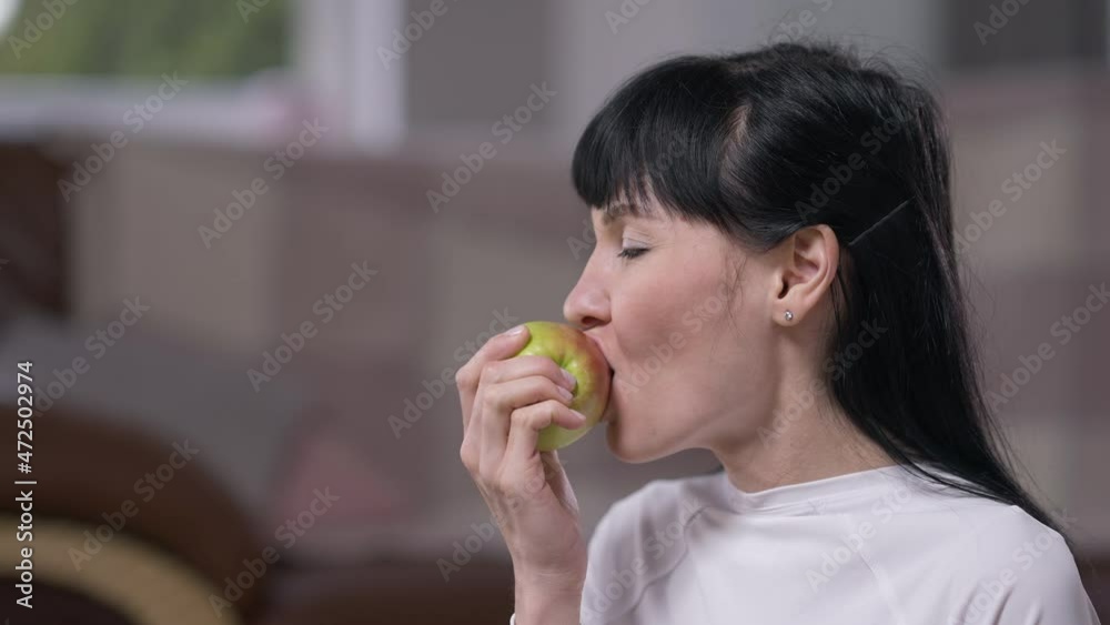 Side view slim woman with black hair biting chewing vitamin apple enjoying taste in slow motion. Portrait of beautiful Caucasian confident lady eating fruit indoors at home. Healthy lifestyle Side view slim woman with black hair biting chewing vitamin apple enjoying taste in slow motion. Portrait of beautiful Caucasian confident lady eating fruit indoors at home. Healthy lifestyle