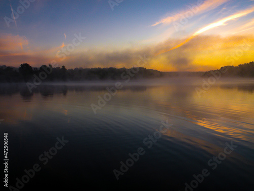 Sunrise over the foggy lake with the reflection of sun. Mist on the water, forest silhouettes and the rays of the rising sun. Beautiful morning landscape with sunrise over river. © MTX