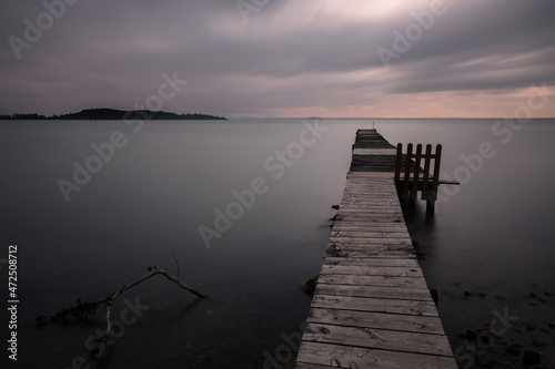 Long exposure view of a pier on a lake at dusk, beneath a dramatic, moody sky
