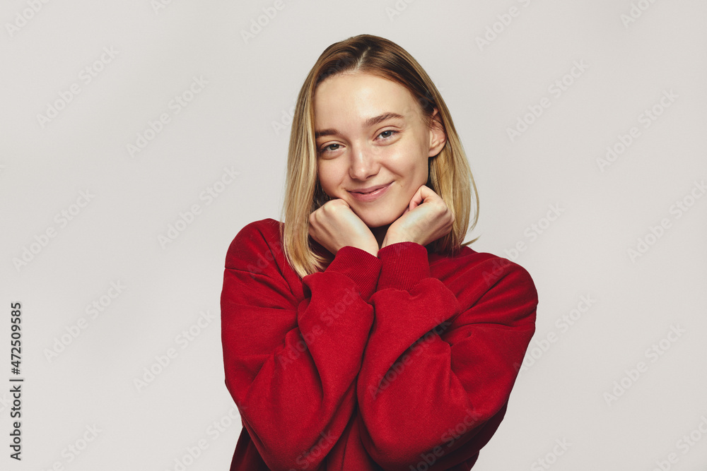 Headshot of pleasant looking cute young woman smiles positively, wears red sweatshirt holds chin with both hands, isolated over white background