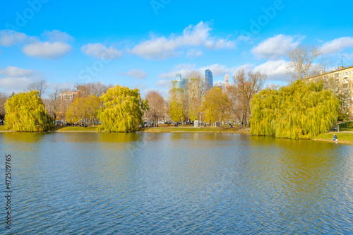 Scenic view of the pond on a sunny autumn day against the backdrop of city buildings. Beautiful landscape with yellow trees on the shore of the pond and the blue sky on an autumn day in the city.