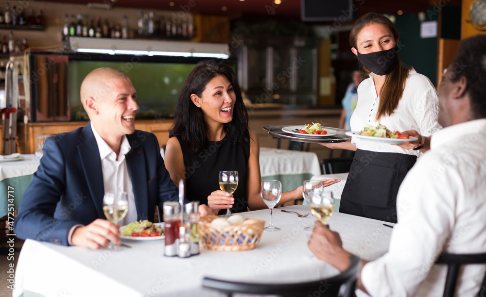 positive female busboy in mask serving order to friends in modern restaurante