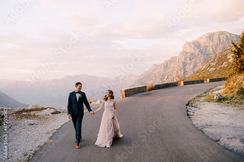 Bride and groom are walking along the asphalt road against the background of mountains