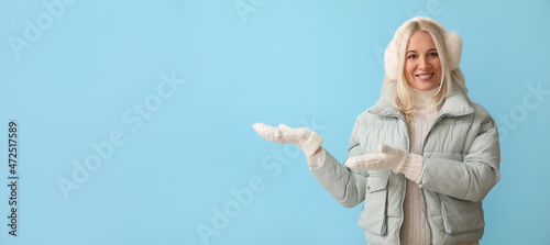 Beautiful mature woman in winter clothes showing something on color background with space for text