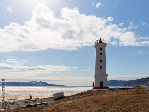 Lighthouse (leading beacon) on the coast of the sea bay. View from the shore to the ice-covered bay. Spring in the Russian Far East. Nagaev Bay, Sea of Okhotsk. Park Lighthouse in the city of Magadan.