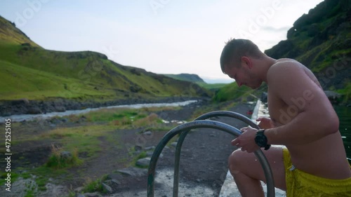 Tourist Climbing Out The Old Swimming Pool Of Seljavallalaug Beside The Narrow River In Iceland. wide shot photo