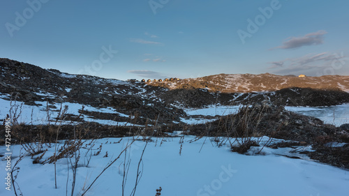 The houses of the tourist base, illuminated by the sun, stand on a snow-covered hill against the blue sky. In the foreground are snowdrifts and dry grass. Siberia.