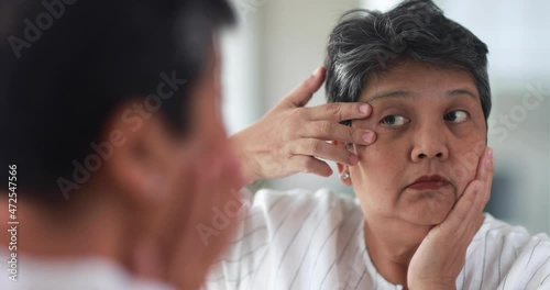 Old Asian woman feels stressed and serious face when watching wrinkle and aging sign around eyes and face in mirror. Elderly lady worrying about beauty. Concept of skin problems in senior people.