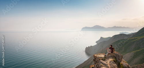 Obraz na płótnie A young Asian woman with a backpack hiking in the summer sits on top of a montage and looks out over a beautiful sea bay landscape