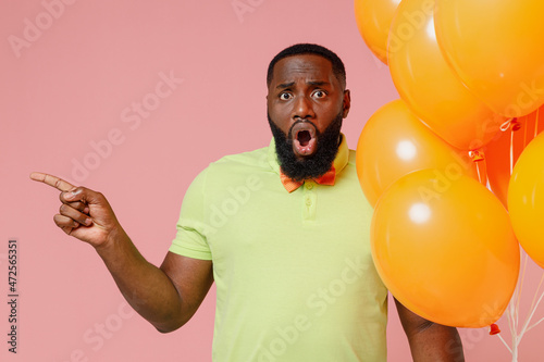 Young disappointed shocked black gay man in green t-shirt bow tie hold bunch air inflated helium balloons celebrating birthday party point index finger aside isolated on plain pastel pink background © ViDi Studio