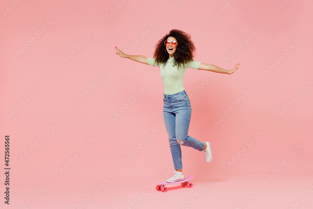 Full size body length happy fun excited bright young curly latin woman 20s wear casual clothes sunglasses skate on board spreading hands isolated on plain pastel light pink background studio portrait