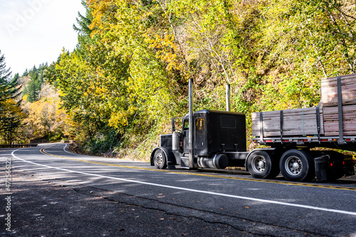 Black classic American bonnet big rig semi truck transporting fastened lamber on flat bed semi trailer running on the winding road with forest on the hill photo