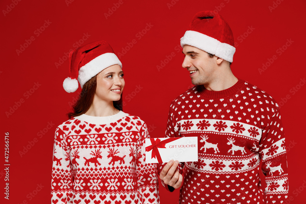 Young fun couple friends two man woman in sweater hat hold gift store certificate coupon voucher card isolated on plain red background. Happy New Year 2022 celebration merry ho x-mas holiday concept.